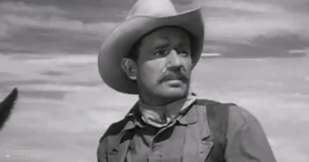 Middle-aged man wearing a wide-brimmed cowboy hat, dark shirt with bandana around neck, set against an open sky suggesting late afternoon or early evening in a desert or plain environment.