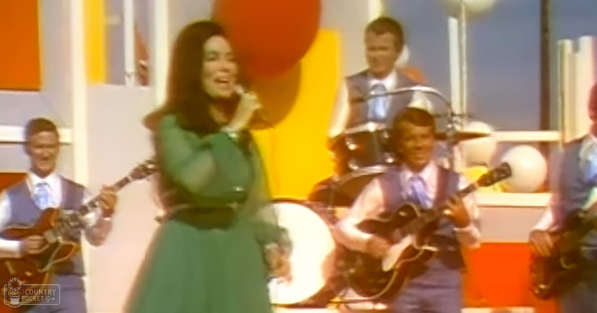 A young adult female performer in a long green dress stands at the forefront of a stage during a mid-20th century performance with other musicians dressed similarly, set against a vibrant orange and blue backdrop.