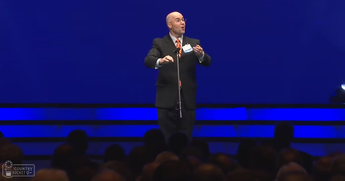 An adult male giving a speech at an indoor event, wearing a dark suit with an orange and black patterned tie, standing on stage under blue lighting.