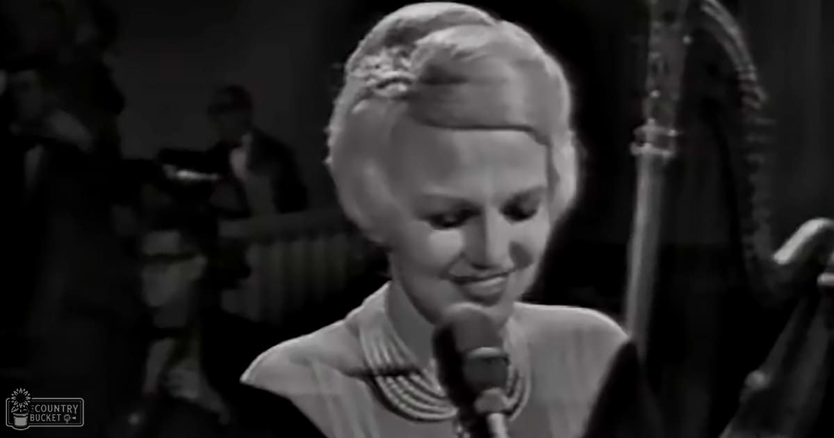 A middle-aged woman in a light-colored garment with floral or abstract patterns stands indoors under artificial lighting during what appears to be an evening event, possibly a performance or recording session.