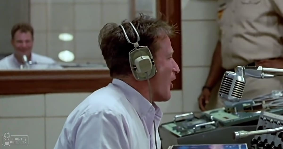A young adult male in a control room, wearing a white button-up shirt and large over-ear headphones from the 1960s or 1970s.