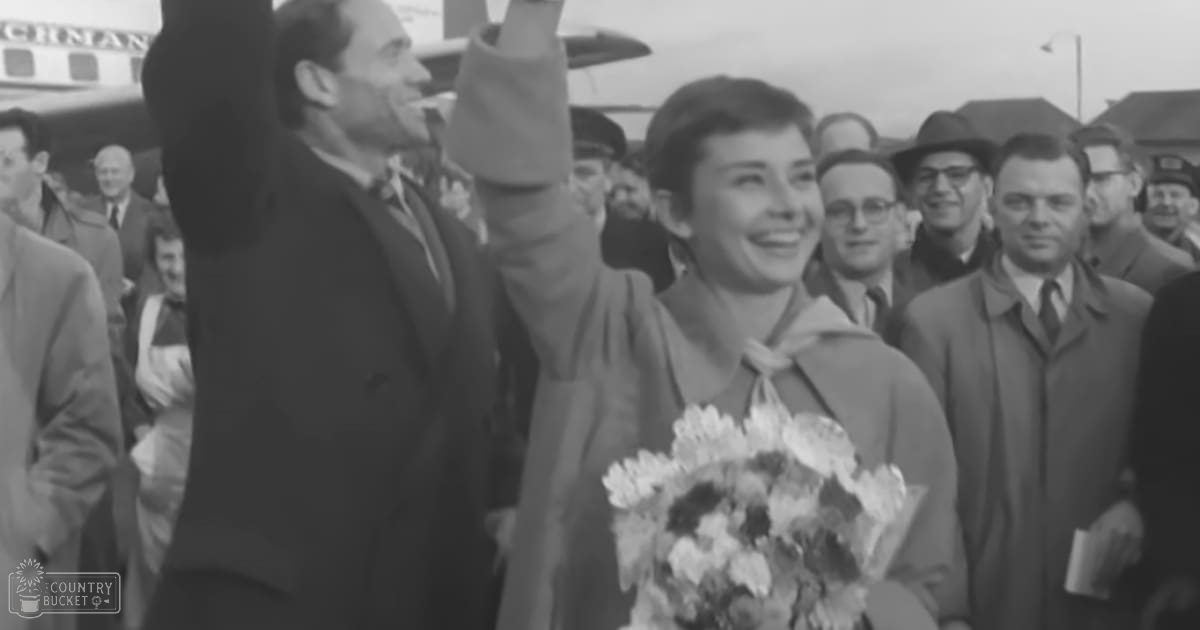 A woman middle-aged with a cheerful expression, holding flowers at an airport setting with other adults dressed formally in the background.