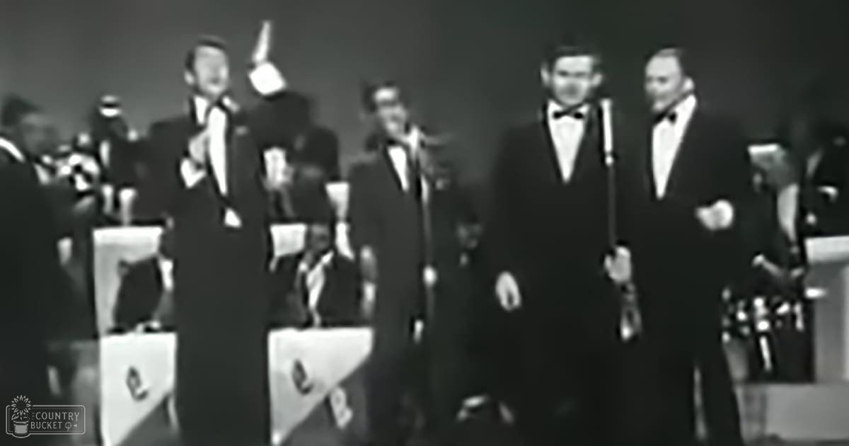 Musical performance scene with multiple adult male singers in dark suits from mid-20th century, indoors under artificial lighting during an evening event.