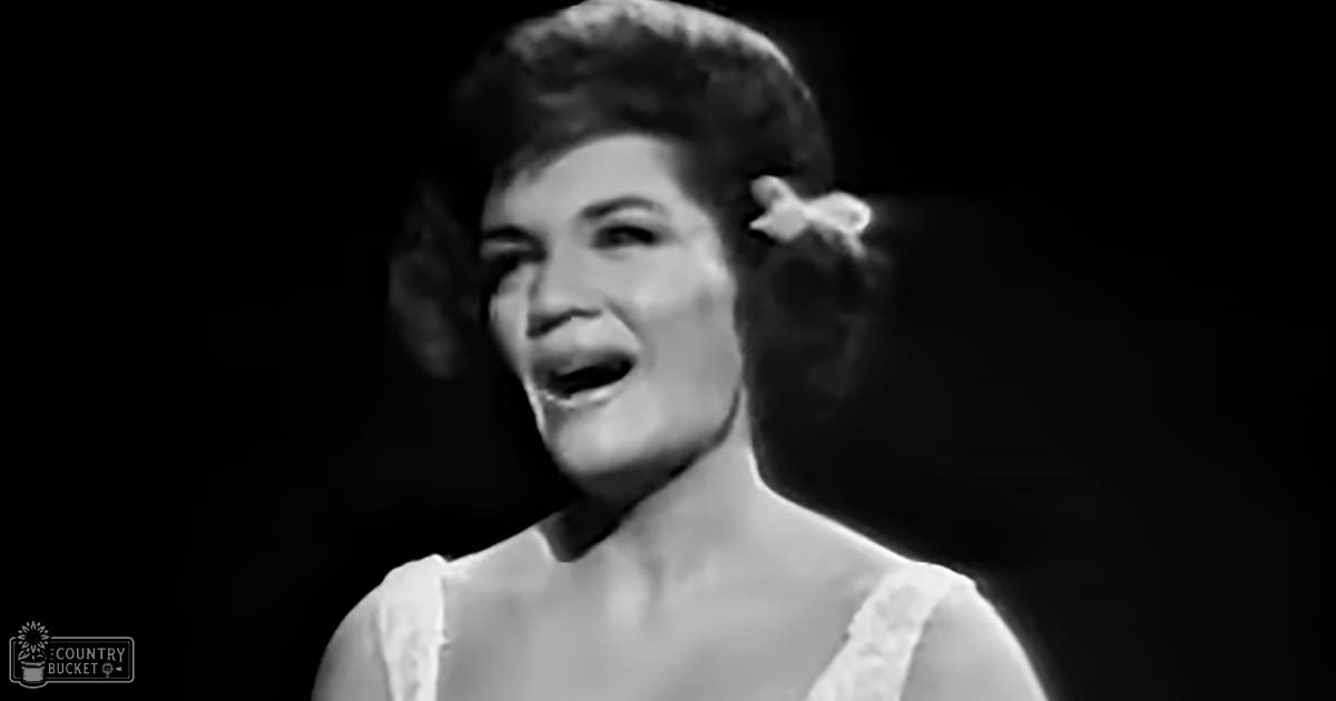 A woman singing on stage wearing a white dress with lace detailing in an indoor performance setting under spotlight lighting.