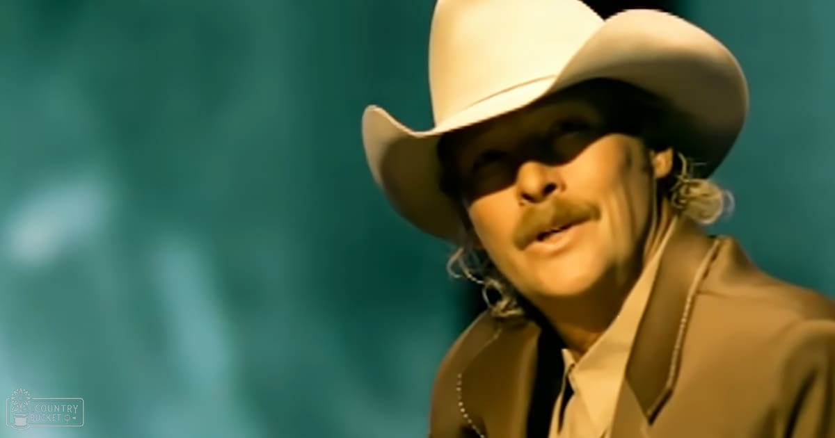 A man in an indoor setting, wearing a white cowboy hat with dark band, beige suit jacket, and white shirt; teal or turquoise lighting.