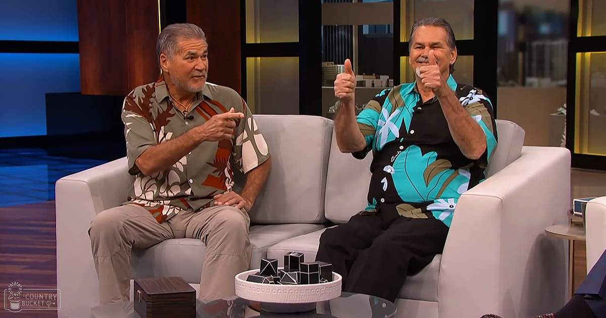 Two older adult males seated on a white couch in a modern studio with dark panels and soft lighting, wearing patterned shirts and beige pants.