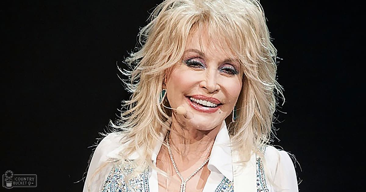 A middle-aged adult female wearing a white sequined blouse with long sleeves, loose silhouette, and V-neck style, accessorized with earrings and a necklace, stands in an indoor setting likely at a stage for a live performance.