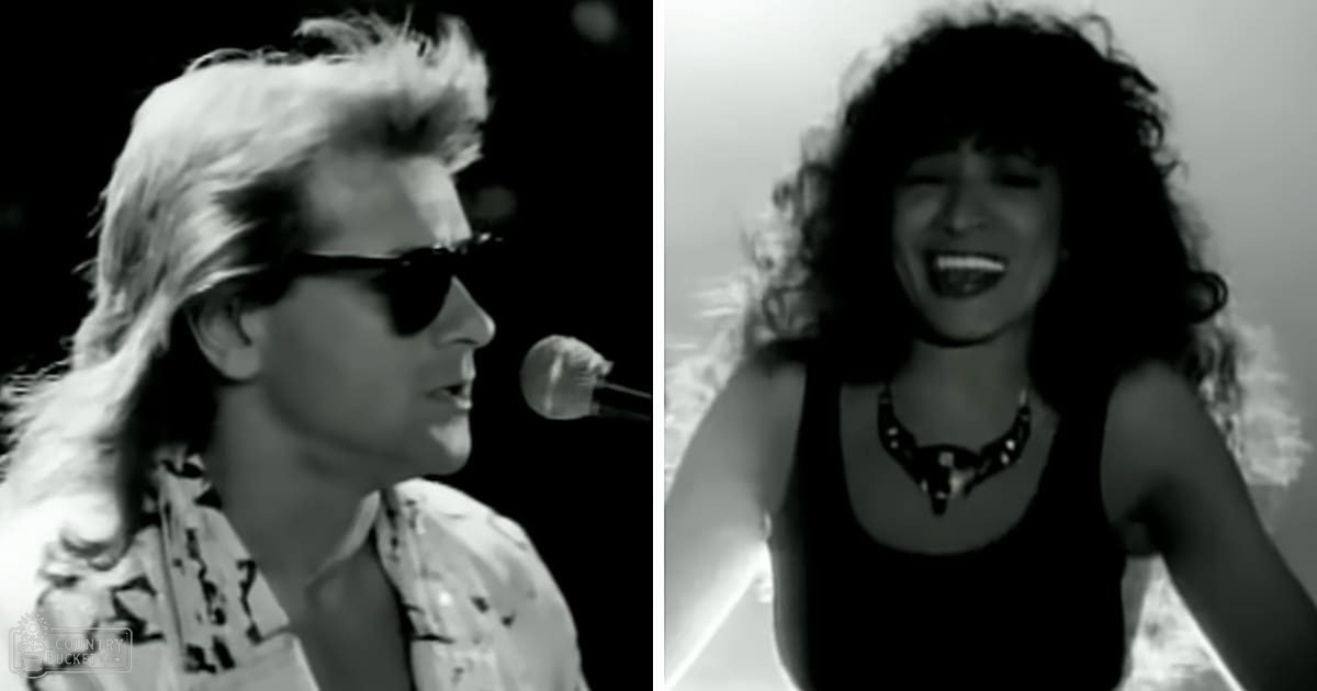 A musical performance scene with two young adults, one male wearing a light-colored patterned shirt and sunglasses, the other female in a dark sleeveless top with a low neckline and a necklace, set against a stage backdrop with professional lighting and smoke or haze.