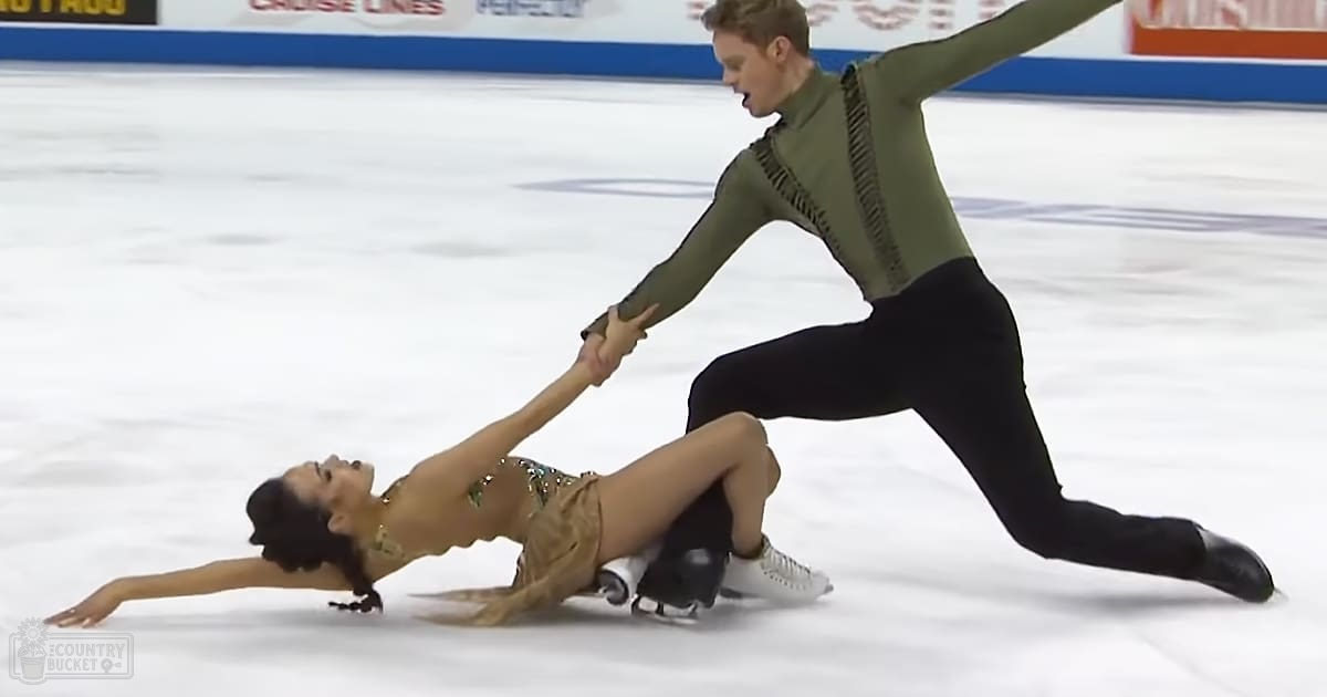A young adult female and male figure skating pair in an indoor rink wearing elegant dresses and athletic attire under bright artificial lighting.