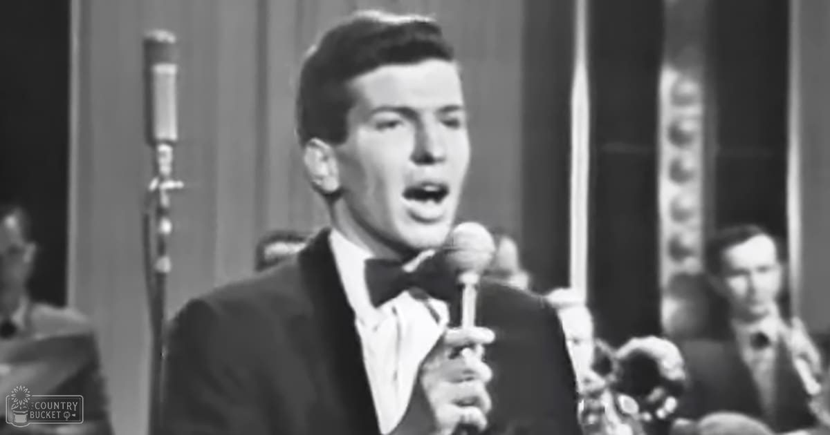 A young adult male singer wearing a dark suit jacket, white shirt with a bow tie, holding a microphone in what seems to be a studio setting from the mid-20th century.