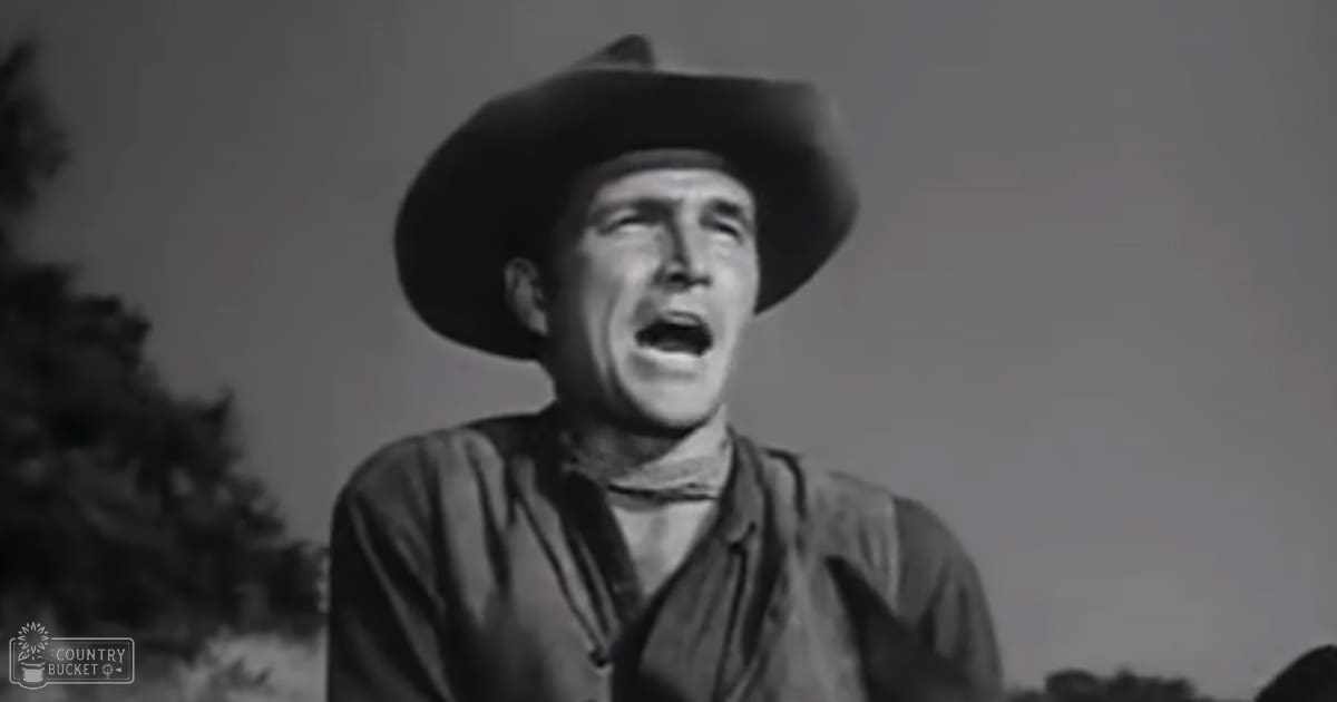 An adult male in a cowboy hat and rugged shirt from what appears to be a Western film, set against a rural outdoor backdrop with trees visible on the left during spring or summer daylight.