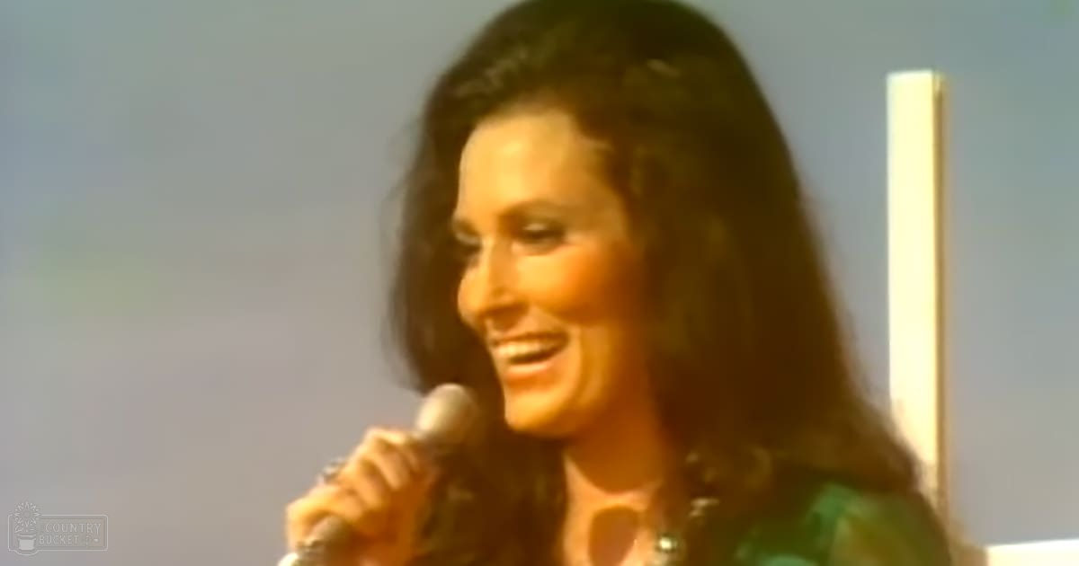 A middle-aged female speaks into a microphone against a simple indoor backdrop, wearing a green patterned V-neck shirt with short sleeves.
