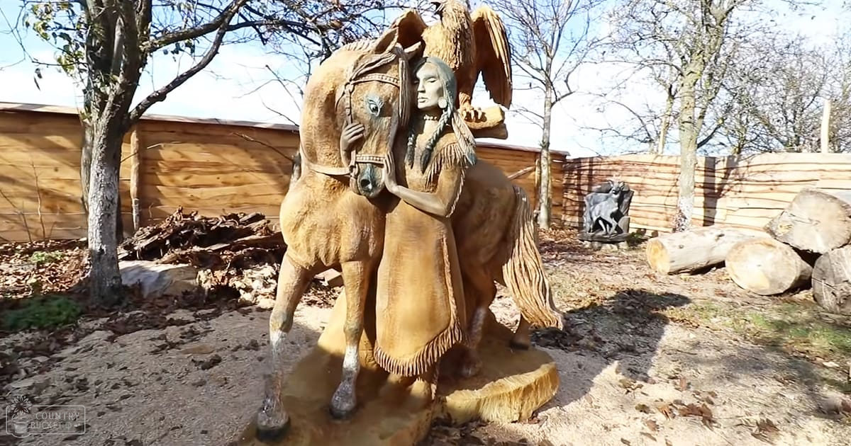 A Native American figure on a horse statue in a rural outdoor setting with wooden fencing and bare trees, suggesting late autumn or winter.