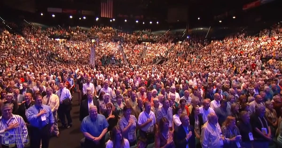 A large crowd of adults gathered inside an indoor arena, likely a convention center, dressed in button-down shirts and casual attire under artificial light.