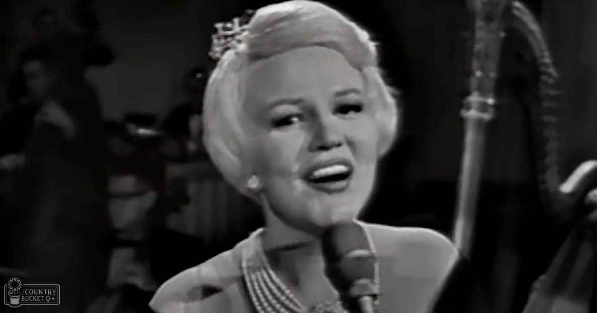 A middle-aged woman singing into a microphone on stage with dimly lit background suggesting an indoor performance venue.