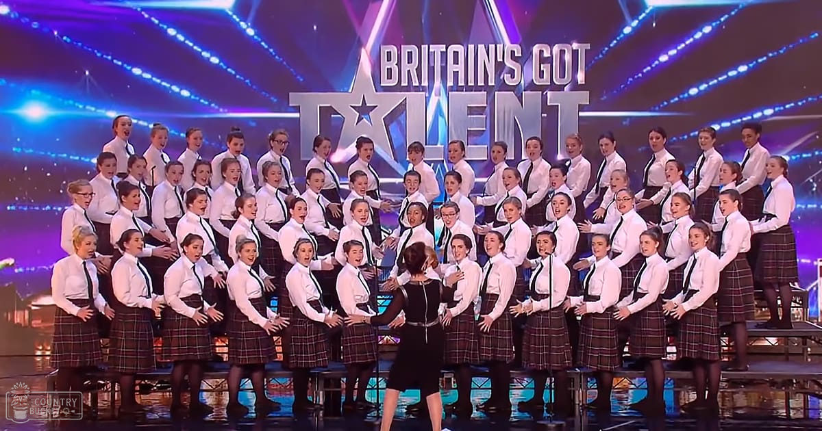 A choir of young female children performing on stage at 'Britain's Got Talent' with blue and purple lighting in a professional talent show setting.