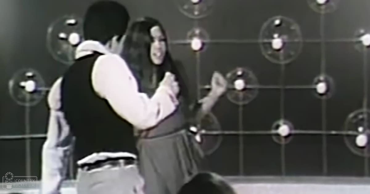 An indoor performance scene with a female performer in a white top and dark skirt, and an adult male in a dark vest and light shirt on a dimly lit stage resembling a mid-20th century concert setting.