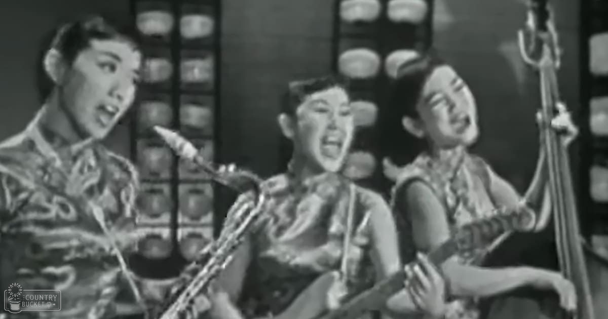 A musical performance scene from an older film or stage production with young adult females wearing intricate patterned garments on a traditional Chinese-themed background.