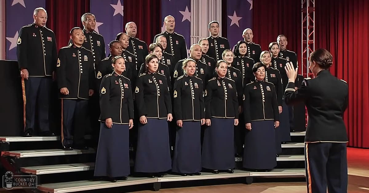 A choir in military-style uniforms performing under bright stage lighting with American flags as backdrop decor, set within a formal indoor performance space.
