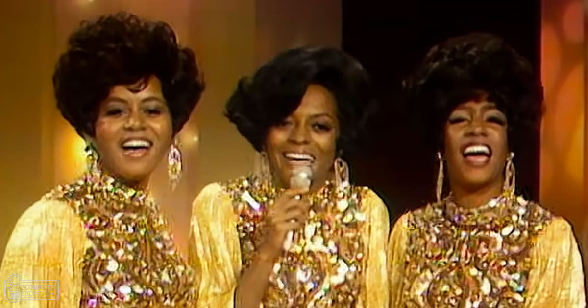 Musical performance scene on stage with three adult females wearing gold sequined tops and large hoop earrings under warm stage lighting, suggesting an evening concert setting.