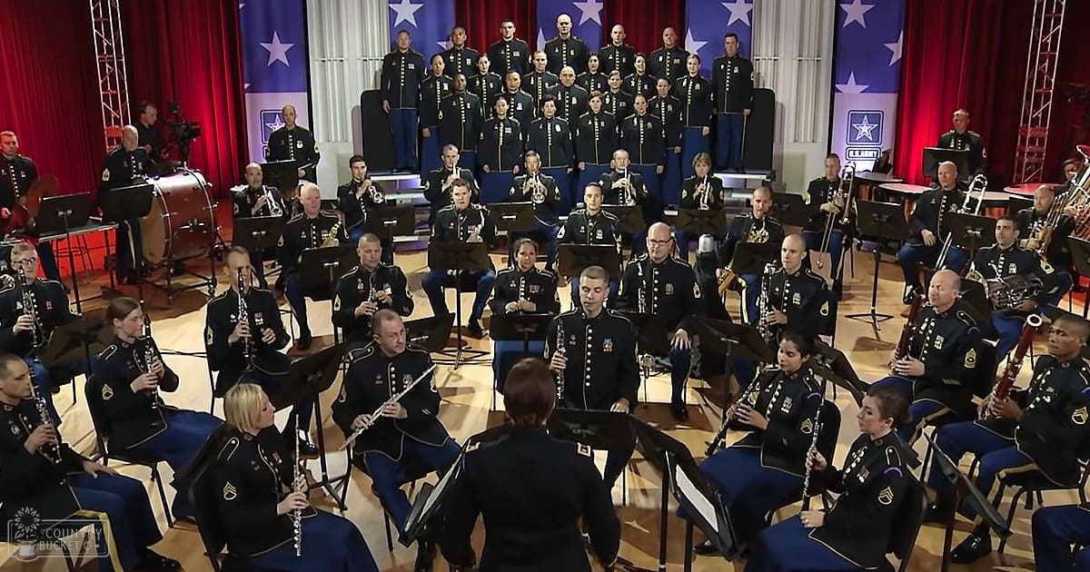 A military band performing in an indoor setting with red curtains, American flags, musical stands, and a conductor holding a baton.