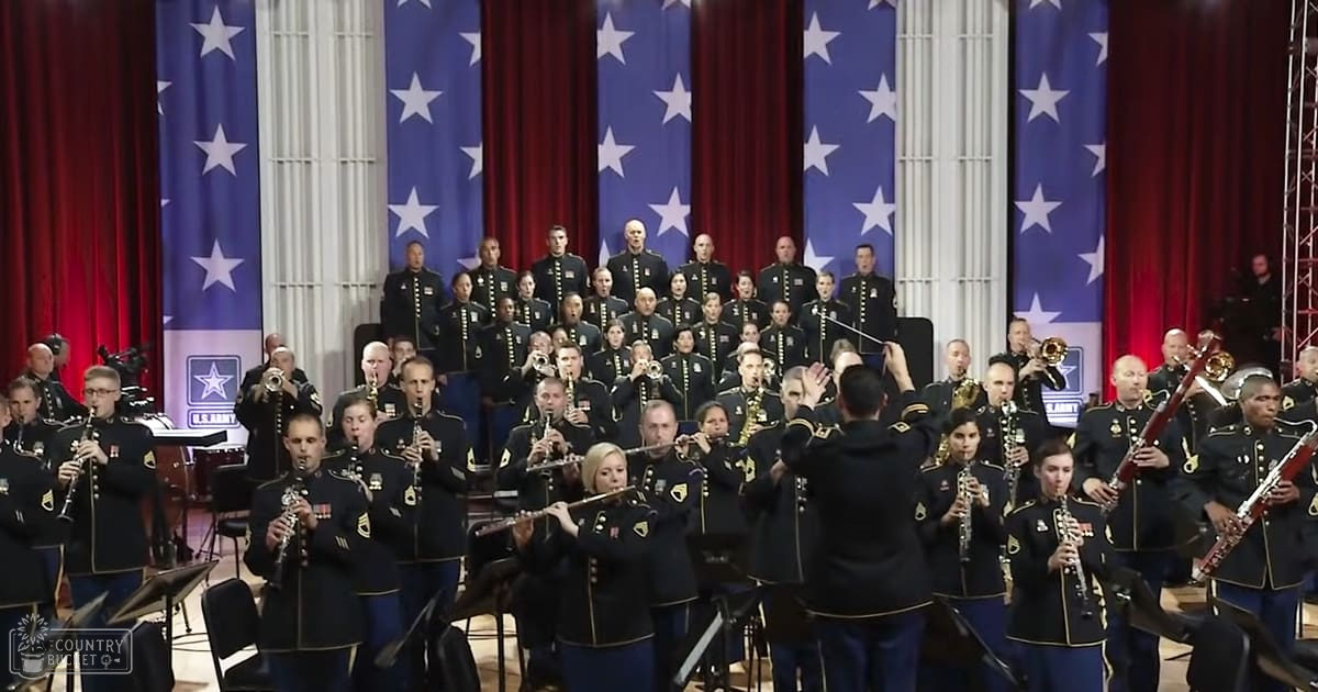 A formal indoor event with a military band performing in front of American flags, with musicians wearing dark blue uniforms and a conductor leading the performance.