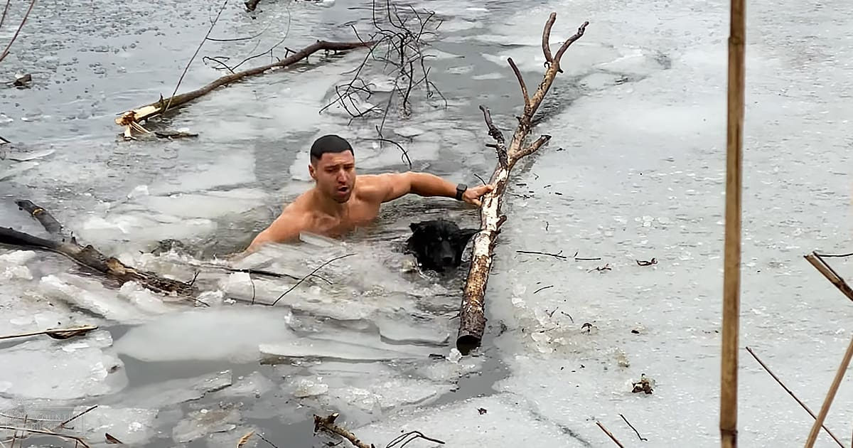 A young adult male wearing swim trunks stands in icy water with broken branches and a partially submerged log, surrounded by ice on what appears to be a lake or river.