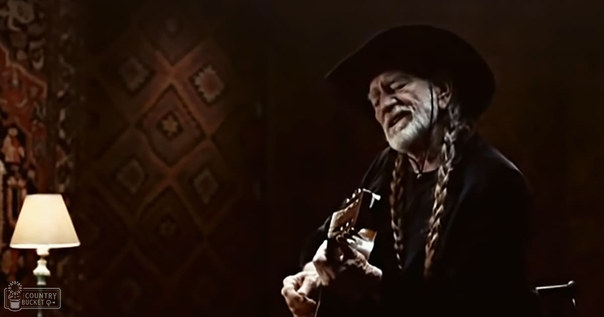 Older adult male playing guitar in a dimly lit indoor setting with geometrically patterned wall decor and a floor lamp.