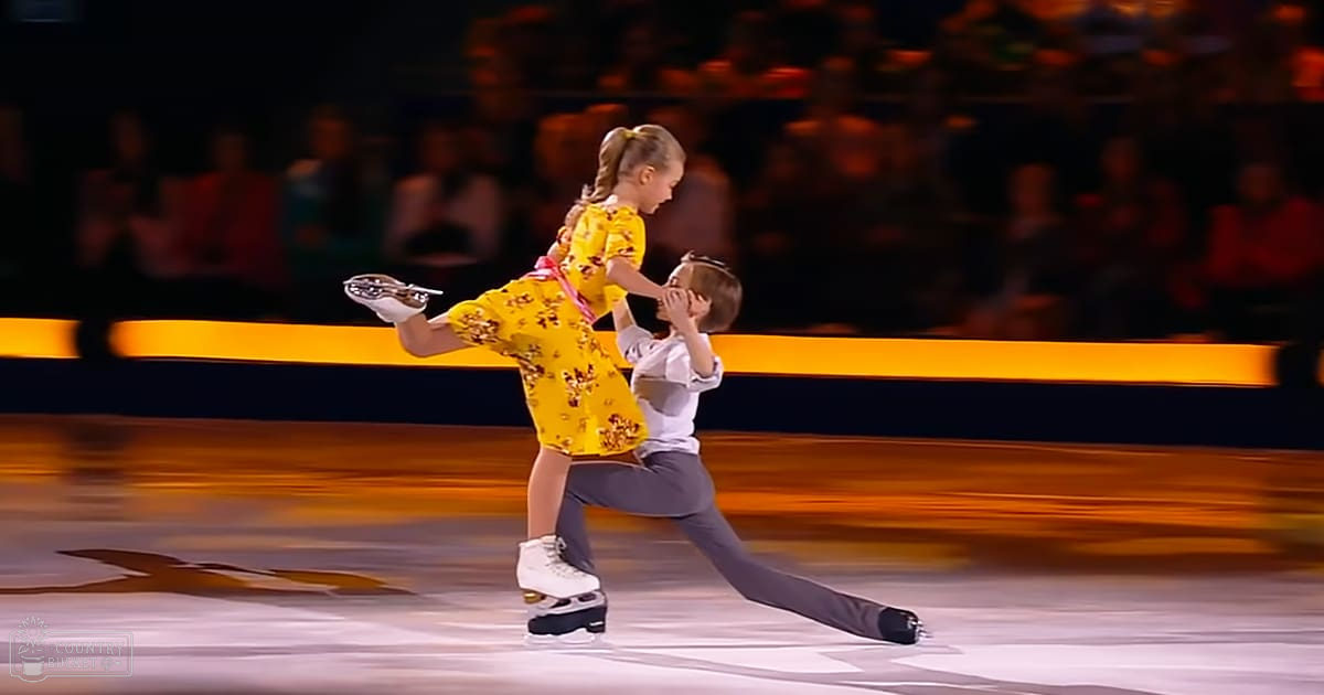 A young female figure skater in a yellow floral dress and red sash performs with her partner, an adult male dressed in gray pants and white shirt, on ice under bright arena lights.