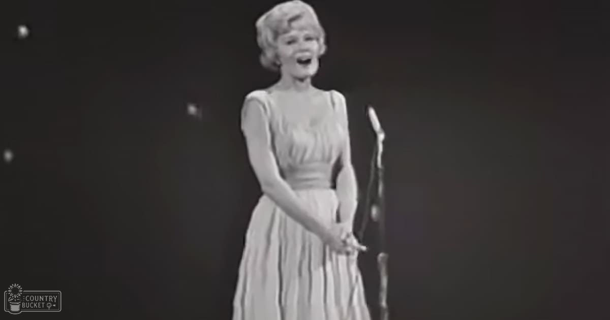 An adult female performer wearing a sleeveless knee-length dress on stage with dark curtains and small white dots in the background.