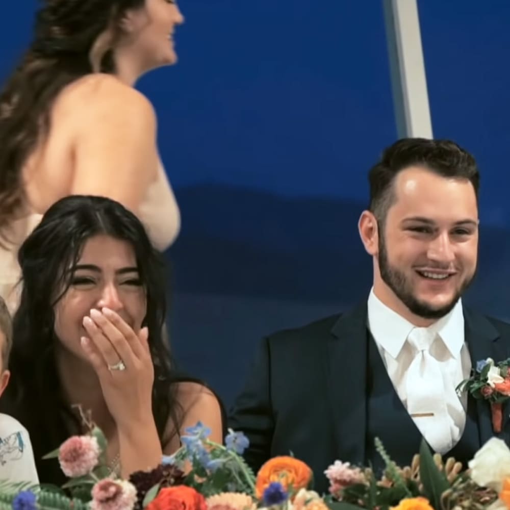 A wedding scene with a young adult female in a sleeveless dress smiling, seated next to an adult male in a formal suit at an indoor venue adorned with flowers and blue walls.