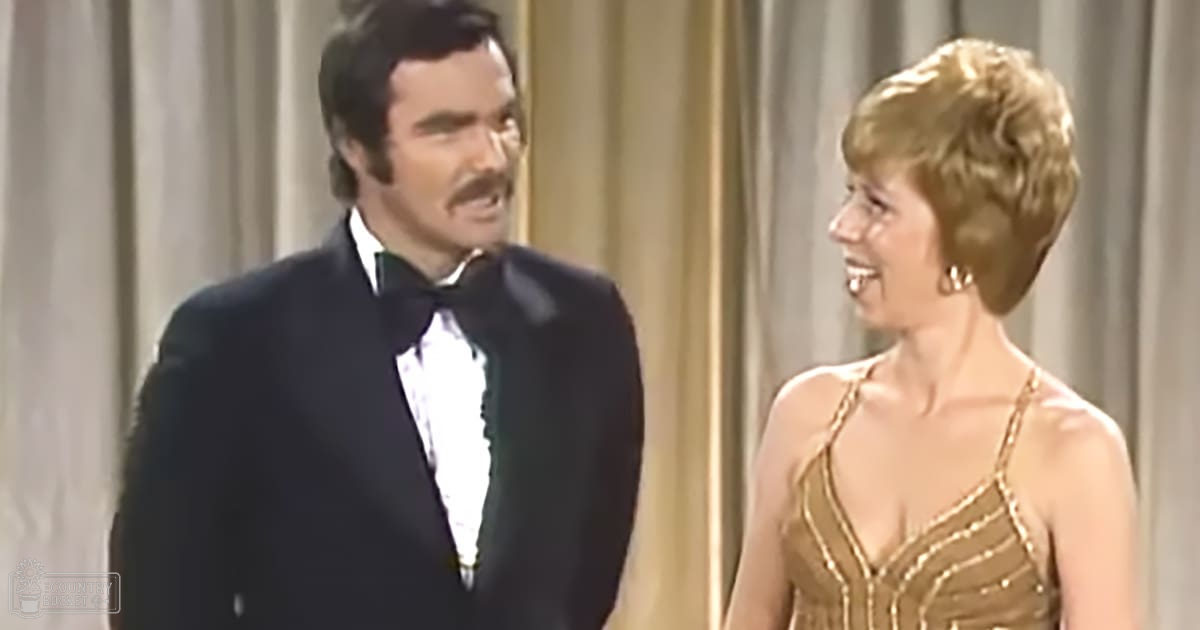 An adult male in a black tuxedo with a white shirt and bow tie, standing indoors at what appears to be an elegant formal gathering or awards ceremony. An adult female in a gold dress with thin straps and sweetheart neckline stands nearby.