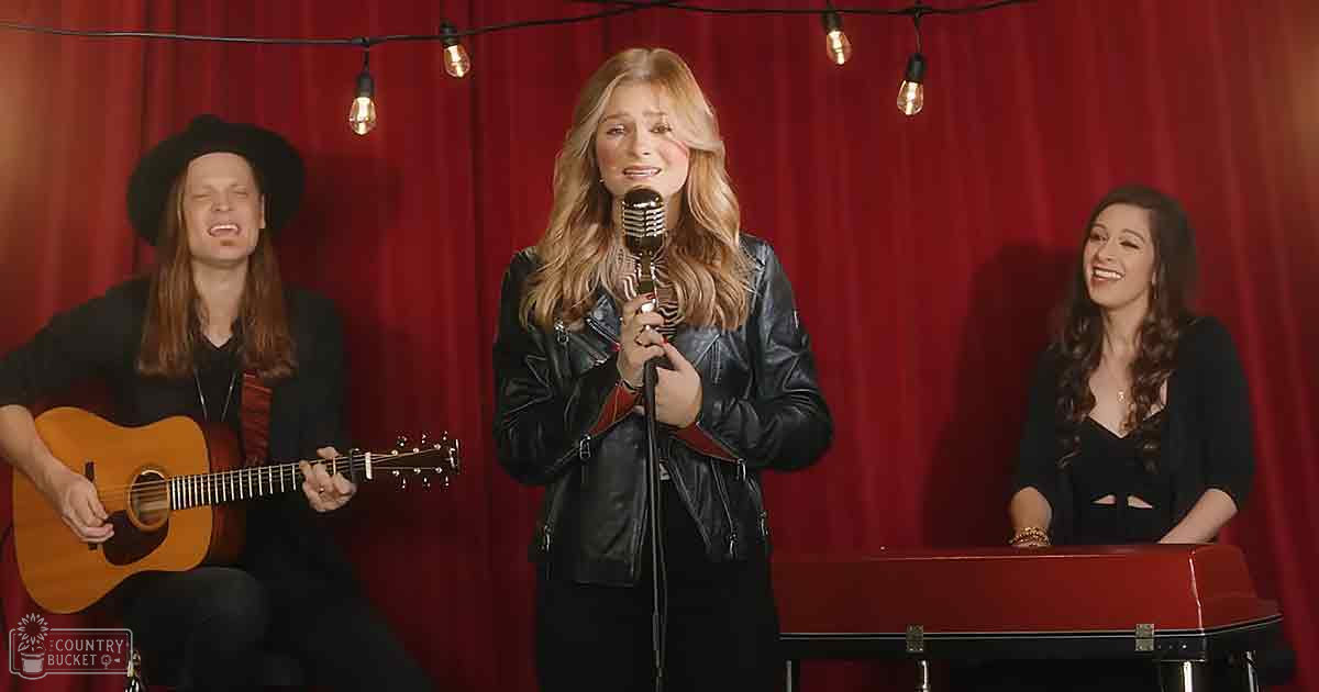 A musical performance with an adult male playing guitar, a female singer holding a microphone, and another female observing, set against a red curtain backdrop with string lights and a piano or keyboard visible.