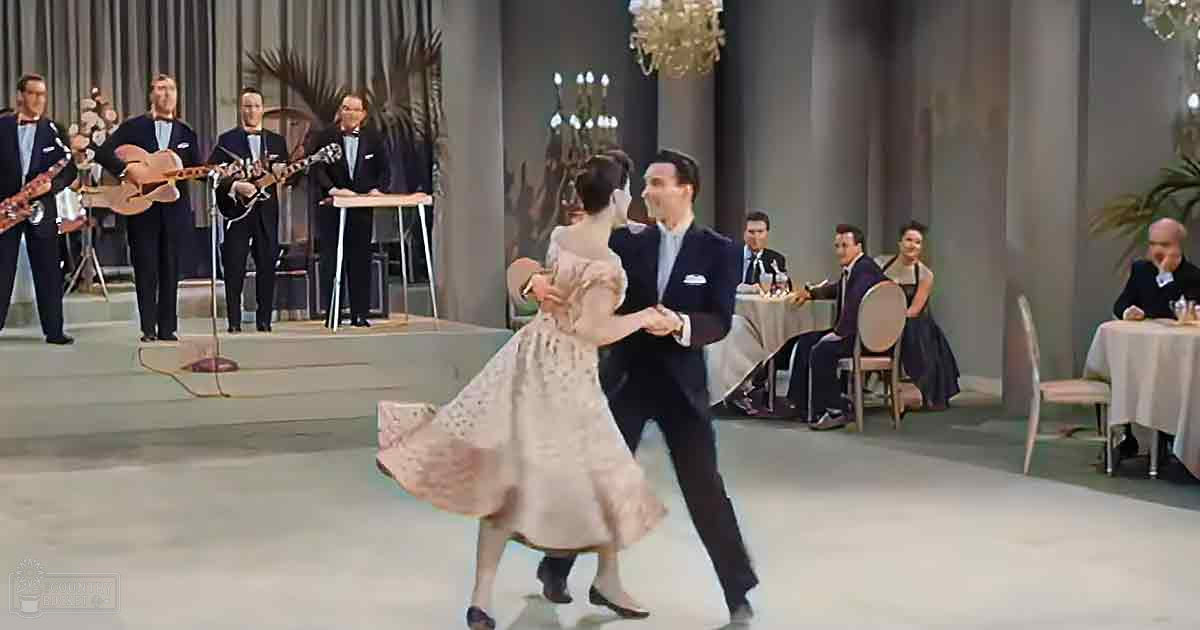 A couple dancing in a formal indoor setting with musicians on stage and chandeliers overhead.