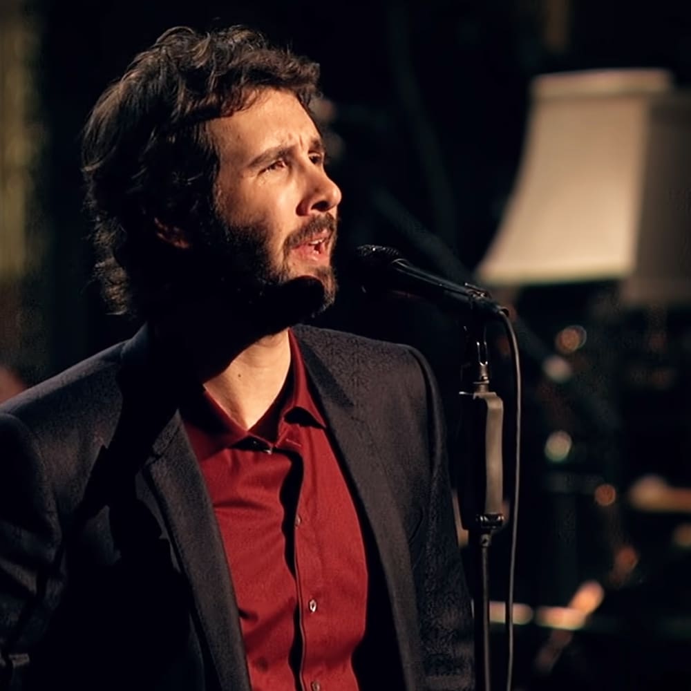 A male adult wearing a dark blazer over a red button-up shirt stands indoors, likely on stage or in a performance space with musical instruments and equipment visible.