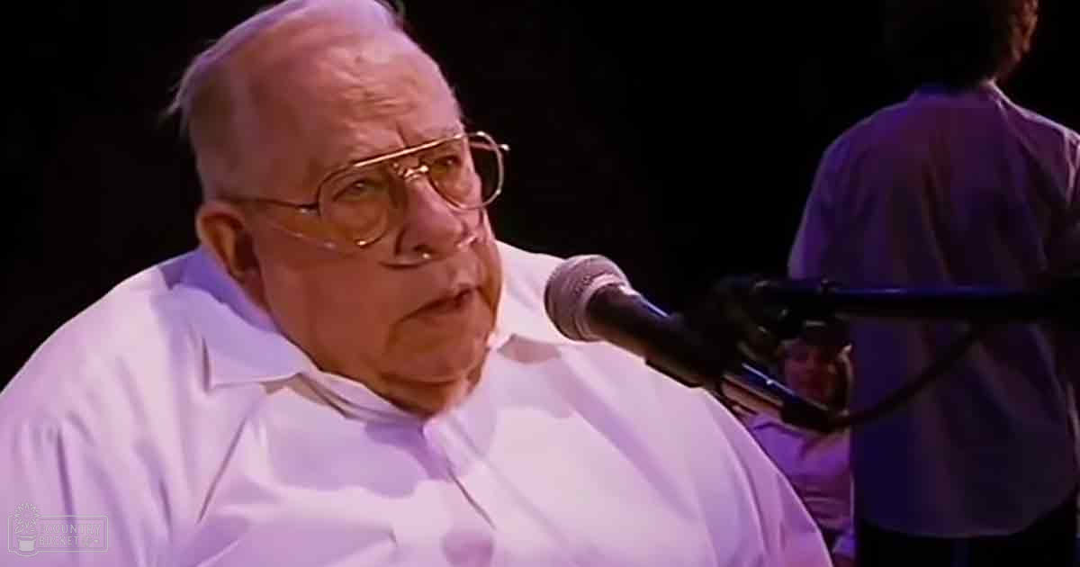 An older adult male performer in a white shirt on a stage with focused lighting during an evening performance, accompanied by another adult male in the background wearing a light-colored shirt and dark pants.