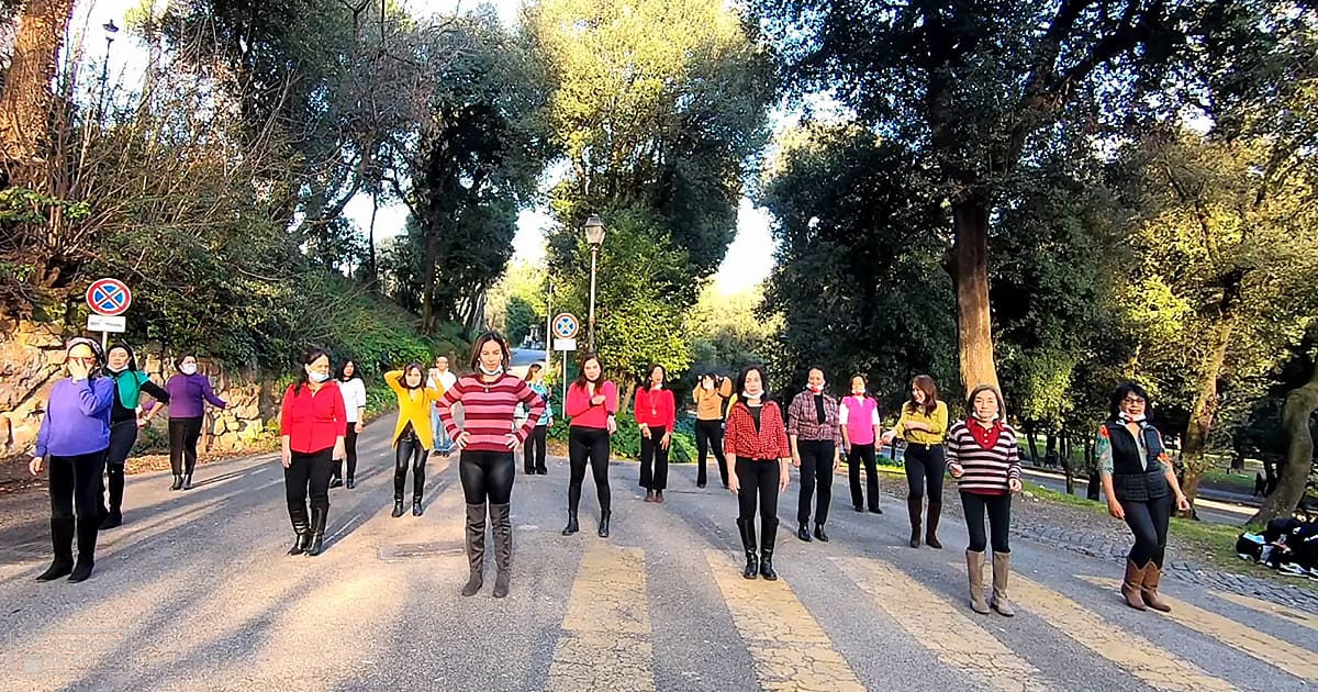 A group of adult women participating in an outdoor activity, possibly dance class or exercise session, on paved paths surrounded by trees with long shadows indicating late afternoon sunlight.