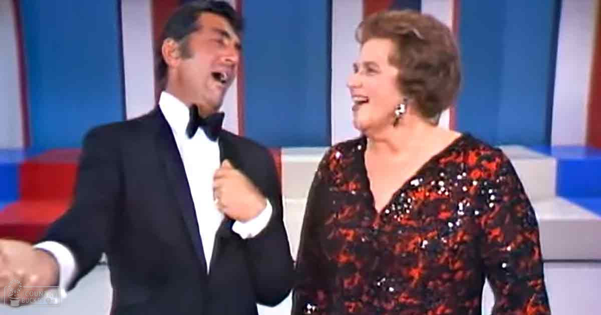 A man in a classic tuxedo and an adult female in a red and black sequined evening gown stand on an indoor stage with American-themed decorations, likely during a formal event.