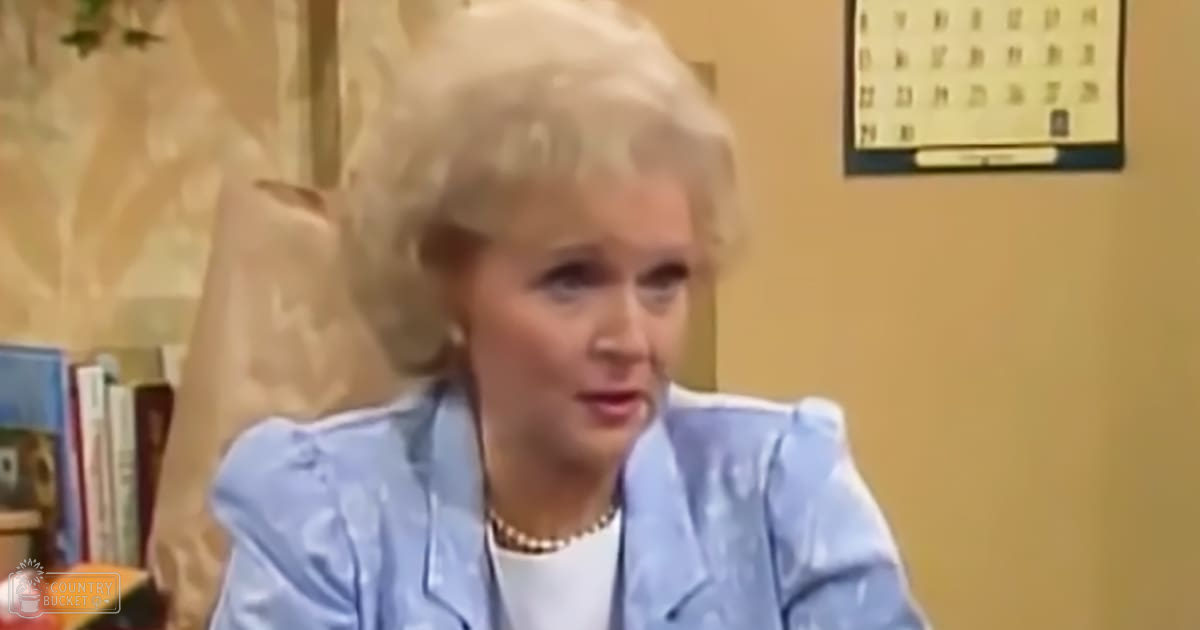 An older adult woman in a light blue, patterned blouse with ruffled shoulders and white undershirt, surrounded by books and floral-patterned decor in an indoor home office setting.
