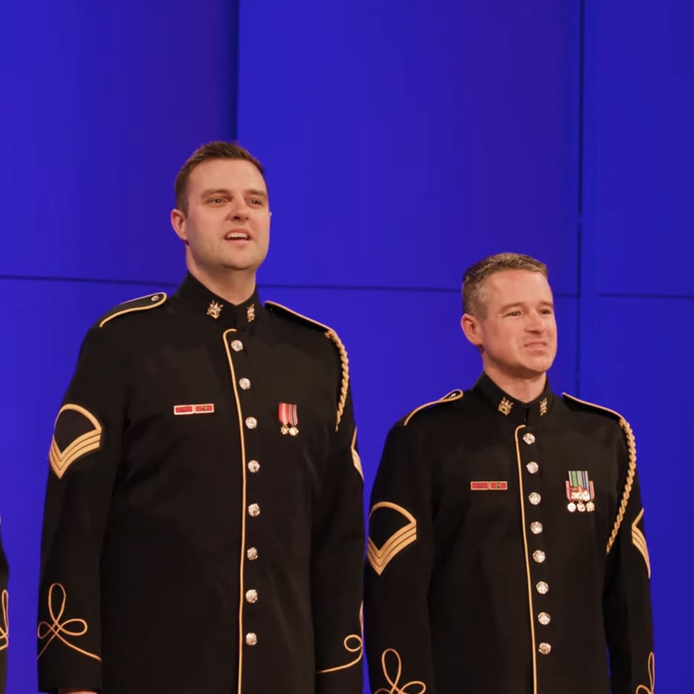 Two adult male individuals dressed in military uniforms with gold braid trim, buttons down the front, red ribbons above chest pockets, and medals near collars, set against a solid blue backdrop indoors under artificial evening lighting.