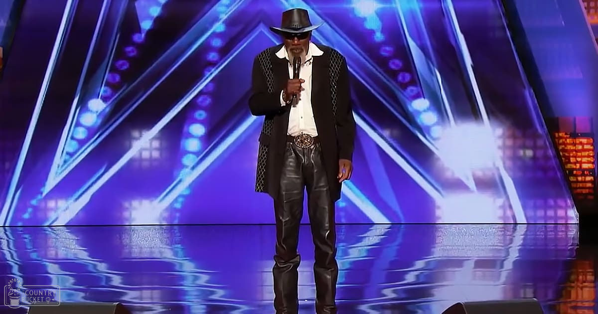An adult male singer wearing a black hat and detailed vest on stage with vibrant purple, blue, and white lighting during an evening event.