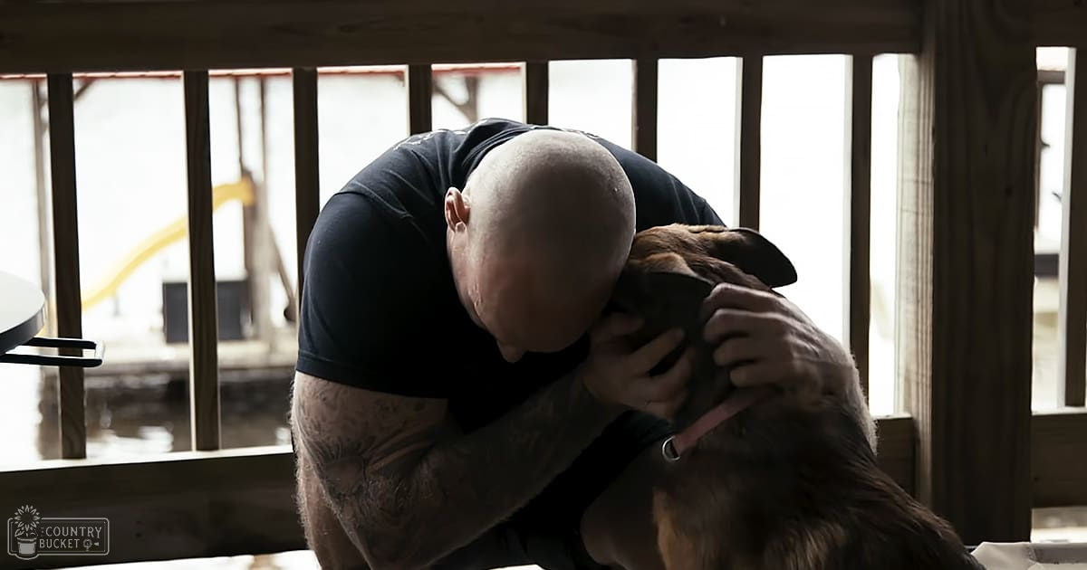 An adult male in a relaxed dark blue T-shirt hugs a brown and black German Shepherd dog inside, with natural light streaming through large windows on a bright spring or summer day.