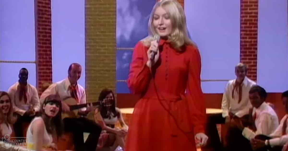 An adult female singer performing on stage at an indoor event, with a red dress and audience watching under stage lighting.