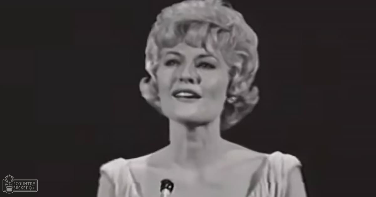 A middle-aged female wearing a light-colored sleeveless top with ruffled shoulders in an indoor setting, holding a microphone.