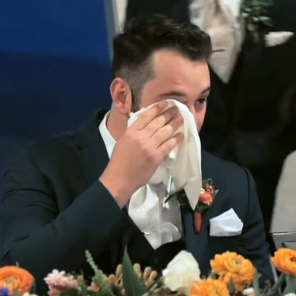 A young adult male in a dark suit with a white shirt and boutonniere, holding a handkerchief at an indoor event with floral decorations.