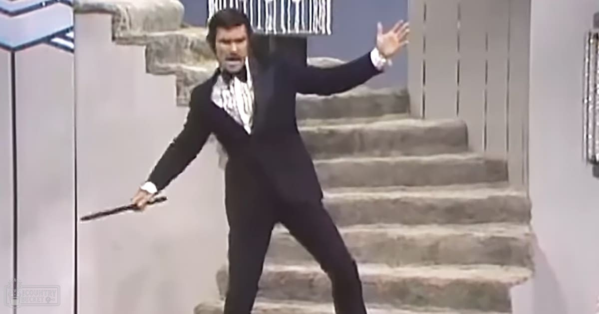 An adult male in a formal black suit with a patterned tie, holding what seems to be a dagger, set outdoors near stone steps during the daytime.