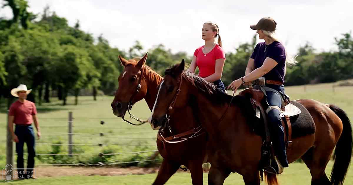 Two adult females riding horses in a rural setting with green grass, trees, and an older male observer standing near a wooden fence. Season appears to be spring or summer.