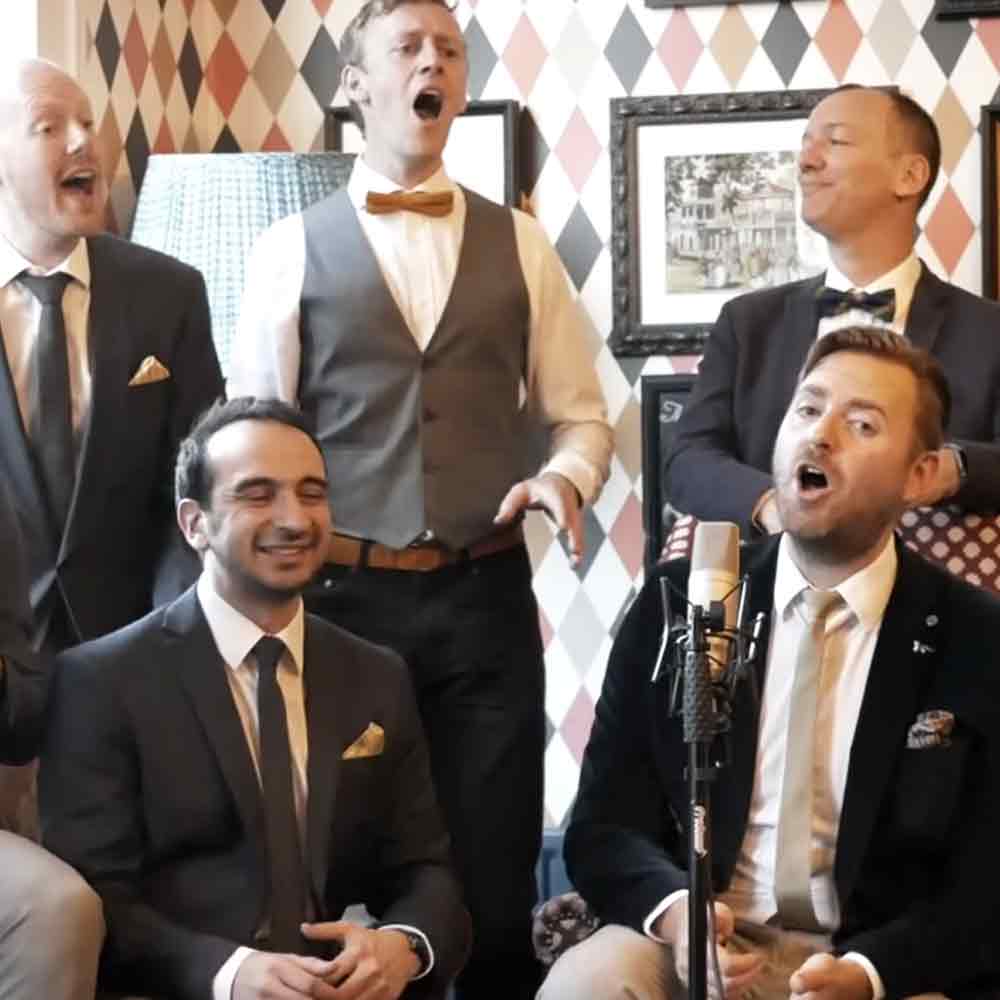 Group of men singing in a bar setting with harlequin-patterned wallpaper and warm indoor lighting.