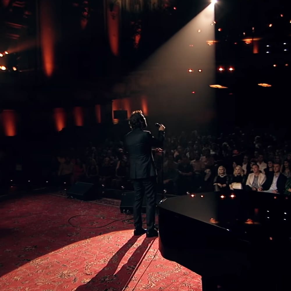 An adult male singer performs on stage under a spotlight at an indoor theater or concert hall with dim lighting and red carpet.