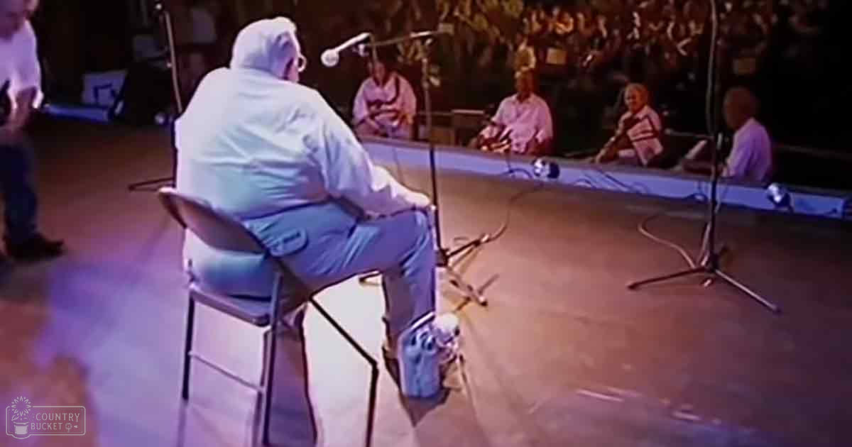 An older adult male in a white hazmat suit seated on stage with microphones around him, suggesting a performance or event at an indoor venue.